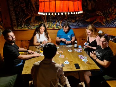 Six individuals gathered around a wooden table in a cozy room, focused on playing a card game with drinks and game materials visible.