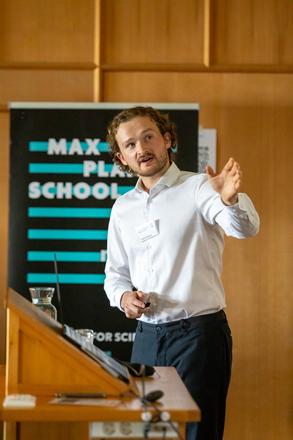 Young man with curly hair and white shirt speaking and gesturing at a wooden podium with a Max Planck School Matter to Life banner in the background.