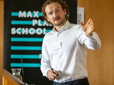Young man with curly hair and white shirt speaking and gesturing at a wooden podium with a Max Planck School Matter to Life banner in the background.