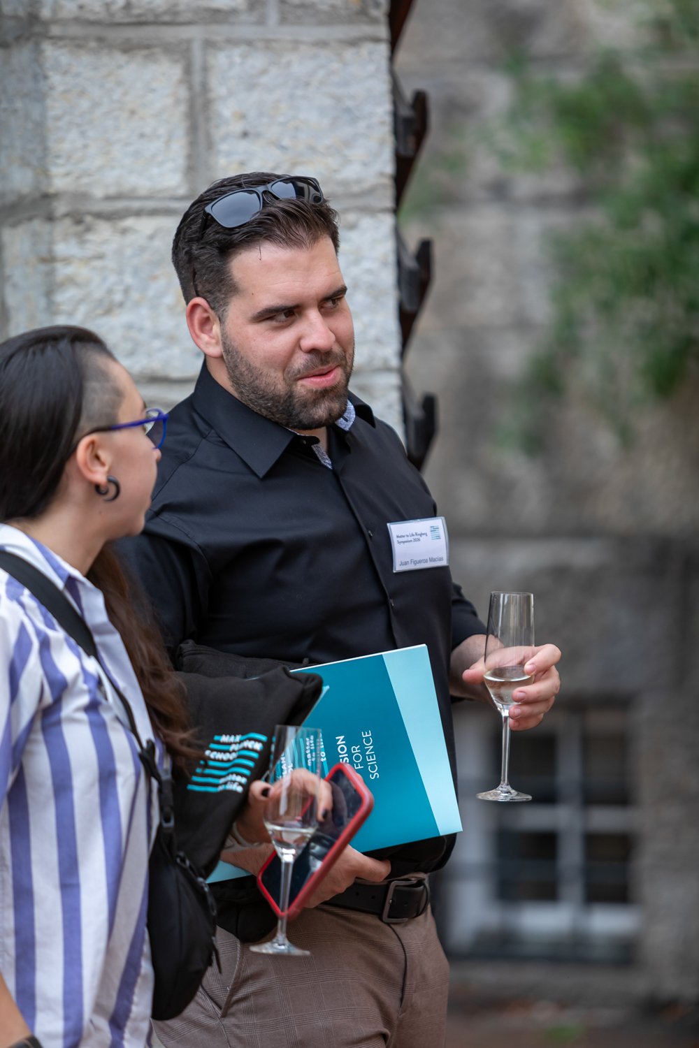 Two individuals talking beside a stone wall, each holding a wine glass; the man wears a name tag and carries a blue folder, while the woman holds a smartphone and wears glasses.