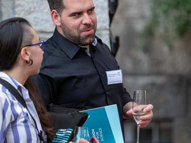Two individuals talking beside a stone wall, each holding a wine glass; the man wears a name tag and carries a blue folder, while the woman holds a smartphone and wears glasses.