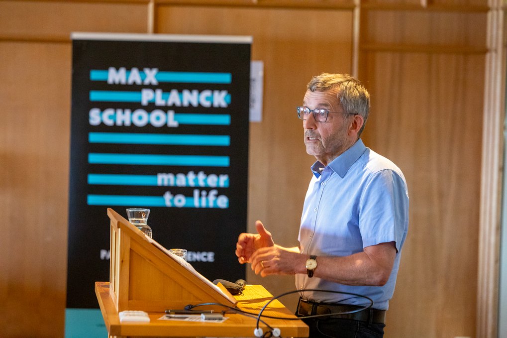Walther Rosenthal wearing glasses and a blue shirt addressing an audience from a podium, with a Max Planck School Matter to Life banner in the background.