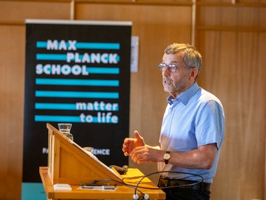 Walther Rosenthal wearing glasses and a blue shirt addressing an audience from a podium, with a Max Planck School Matter to Life banner in the background.