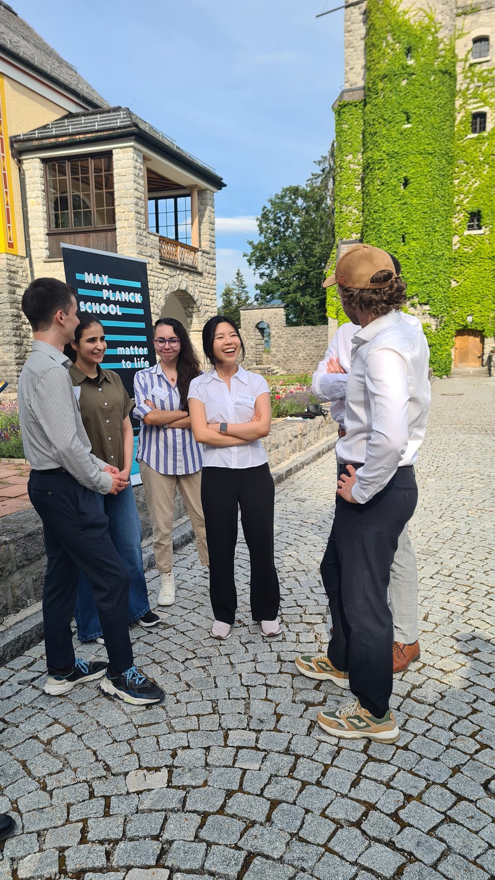 Six individuals engaged in conversation on a cobblestone courtyard near a stone building covered with green ivy and a Max Planck School Matter to Life sign.