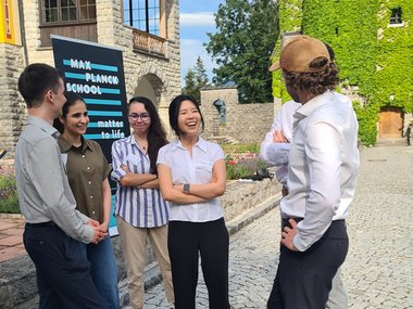 Six individuals engaged in conversation on a cobblestone courtyard near a stone building covered with green ivy and a Max Planck School Matter to Life sign.