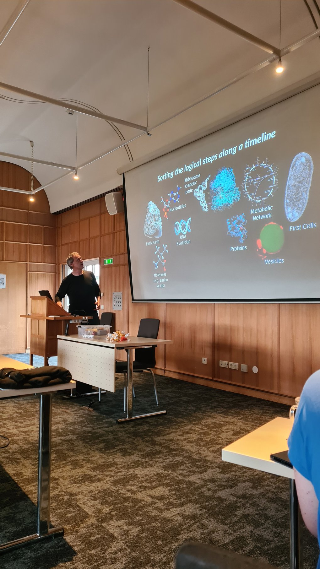 Presenter standing at a podium in a conference room, explaining a projected slide illustrating the logical steps from molecules to first cells.