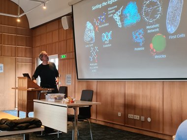 Presenter standing at a podium in a conference room, explaining a projected slide illustrating the logical steps from molecules to first cells.