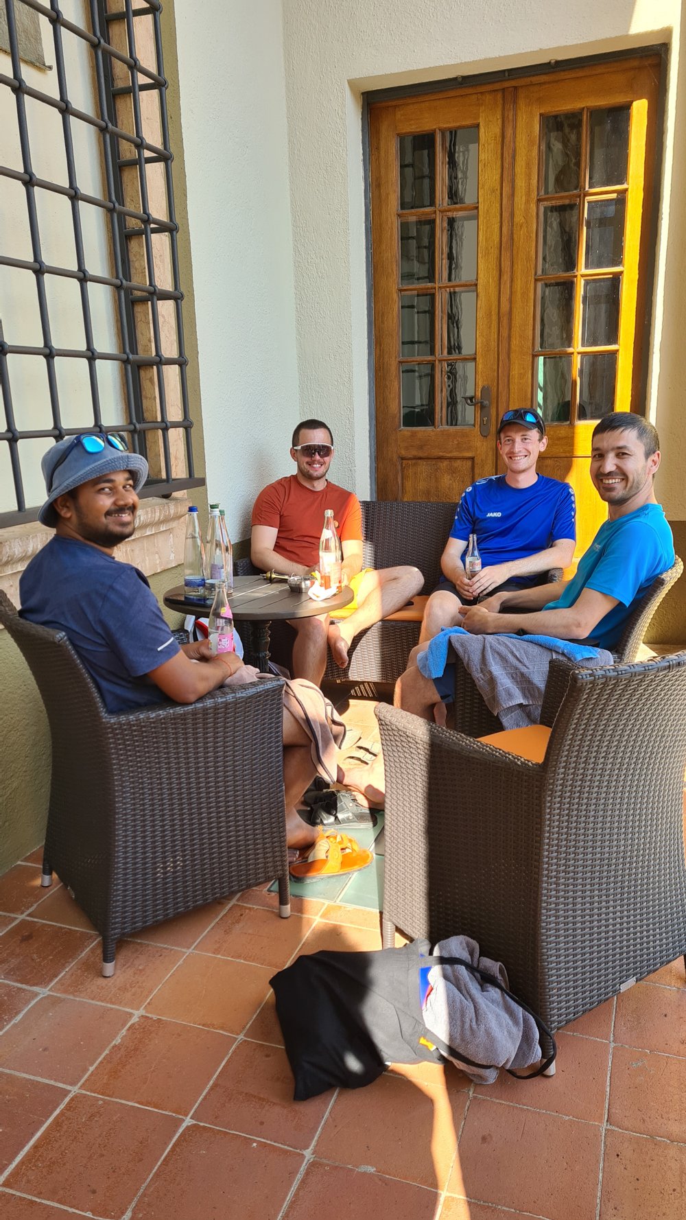 Four men relaxing on wicker chairs around a table with bottles, in front of a wooden door on a sunlit tiled patio.