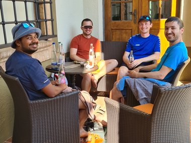 Four men relaxing on wicker chairs around a table with bottles, in front of a wooden door on a sunlit tiled patio.