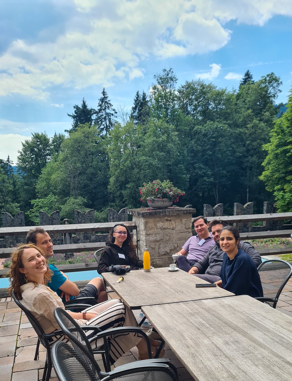 Six people gathered around an outdoor table on a stone-paved patio, with forested scenery and a stone wall adorned with a flowerpot behind them.
