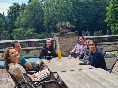 Six people gathered around an outdoor table on a stone-paved patio, with forested scenery and a stone wall adorned with a flowerpot behind them.