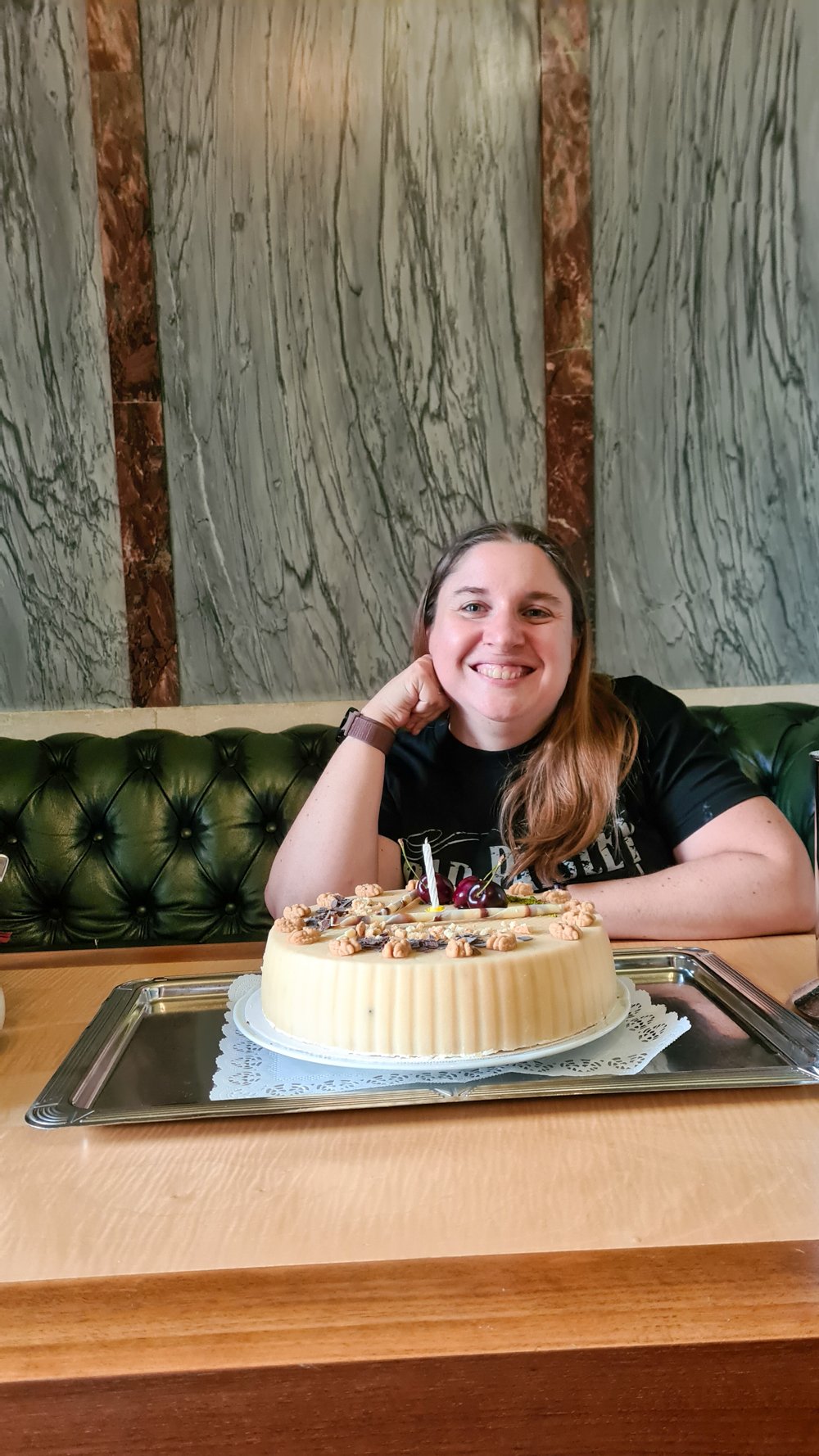 Woman with long hair sitting on a green tufted bench behind a wooden table, with a round cake decorated with nuts and cherries placed on a silver tray in front of her.