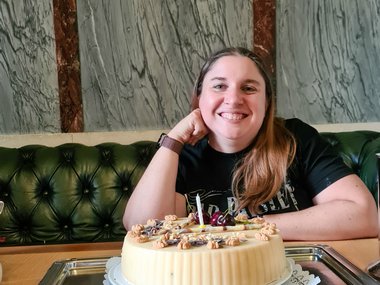 Woman with long hair sitting on a green tufted bench behind a wooden table, with a round cake decorated with nuts and cherries placed on a silver tray in front of her.