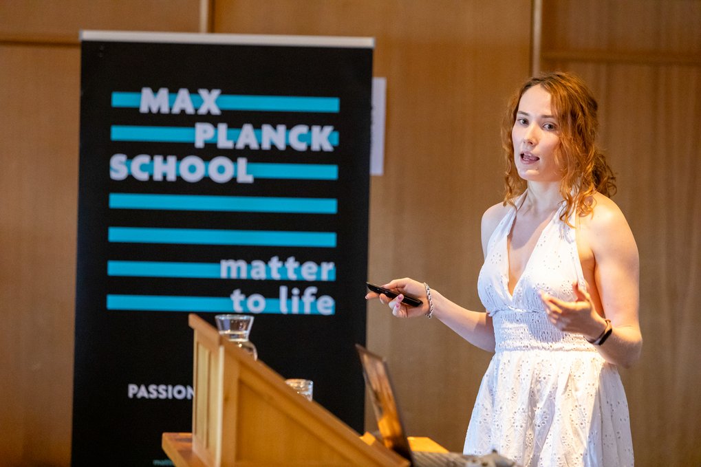 A woman in a white sleeveless dress is delivering a presentation at a wooden podium, holding a remote control, with a Max Planck School Matter to Life banner visible in the background.