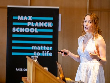 A woman in a white sleeveless dress is delivering a presentation at a wooden podium, holding a remote control, with a Max Planck School Matter to Life banner visible in the background.
