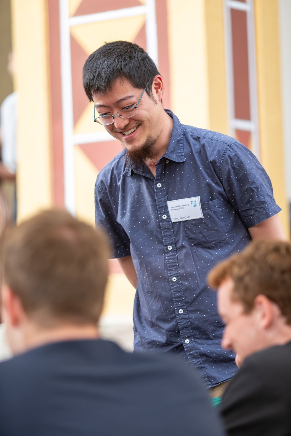 Man with glasses and a short beard, wearing a blue shirt with small white dots, smiling and interacting with two individuals seated in a room with decorative walls.