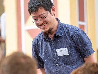 Man with glasses and a short beard, wearing a blue shirt with small white dots, smiling and interacting with two individuals seated in a room with decorative walls.