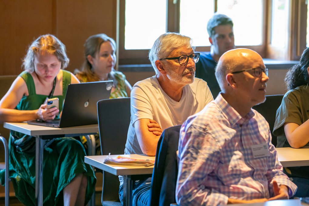 Several adults sitting at desks in a lecture room setting, some using laptops and others listening attentively.
