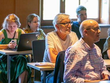 Several adults sitting at desks in a lecture room setting, some using laptops and others listening attentively.