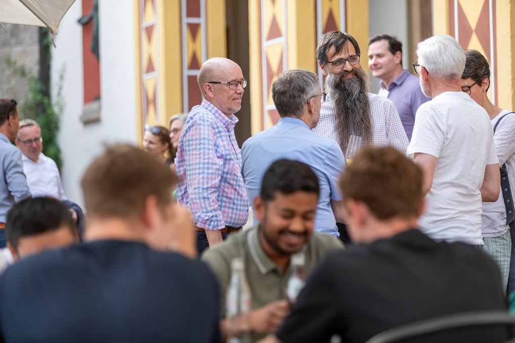 Several individuals engaged in conversation outside, with some sitting at a table and others standing near a building featuring geometric wall designs.