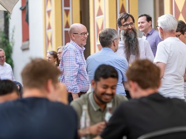 Several individuals engaged in conversation outside, with some sitting at a table and others standing near a building featuring geometric wall designs.