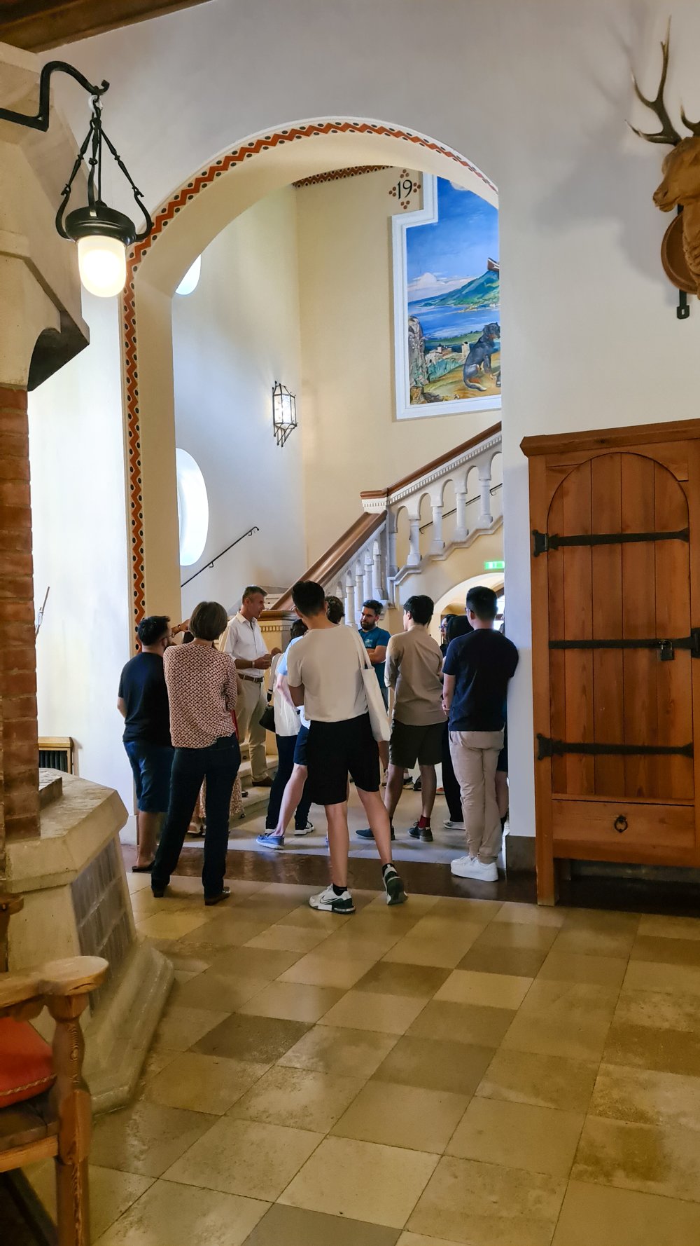Several individuals gathered in a room with tiled floors, an arched doorway, and a staircase adorned with a colorful mural.