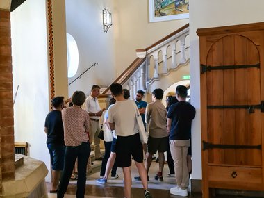 Several individuals gathered in a room with tiled floors, an arched doorway, and a staircase adorned with a colorful mural.