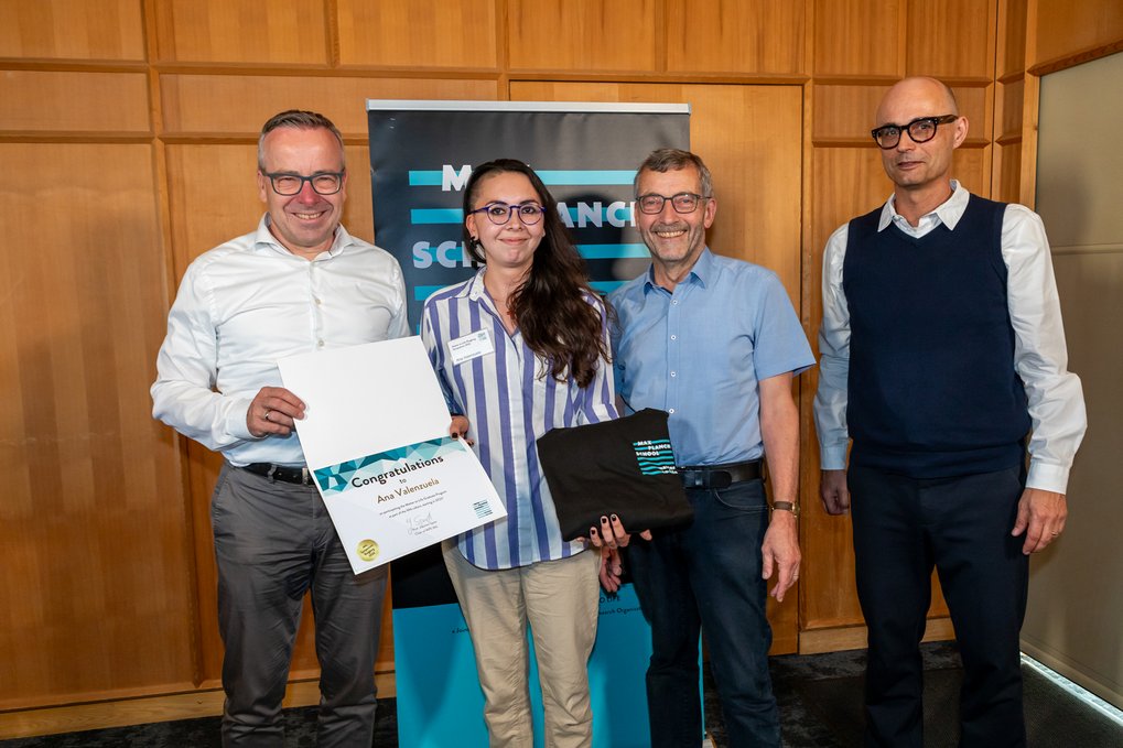 Gunther Friedl, Ana Valenzuela, Walter Rosenthal, and Joachim Spatz are standing side by side in a wood-paneled room; one person is holding a certificate addressed to Ana Valenzuela, and another is holding a black hoodie with the “Matter to Life” logo—they are all smiling at the camera.