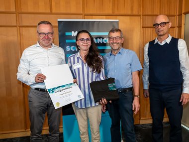 Gunther Friedl, Ana Valenzuela, Walter Rosenthal, and Joachim Spatz are standing side by side in a wood-paneled room; one person is holding a certificate addressed to Ana Valenzuela, and another is holding a black hoodie with the “Matter to Life” logo—they are all smiling at the camera.