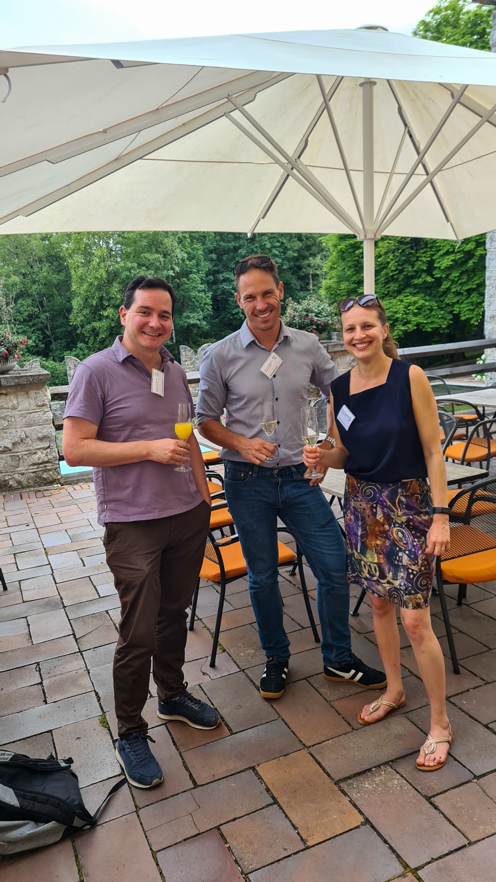 Three people standing under a large white umbrella on a patio holding drinks.