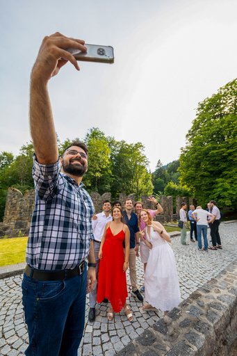 Ringberg Symposium 2026: A candid moment from the 5th cohort's Master's graduation ceremony at Ringberg Castle – marking the transition from the Master's phase to PhD research A diverse group of friends posing for a selfie in a park with trees and stone structures in the background.